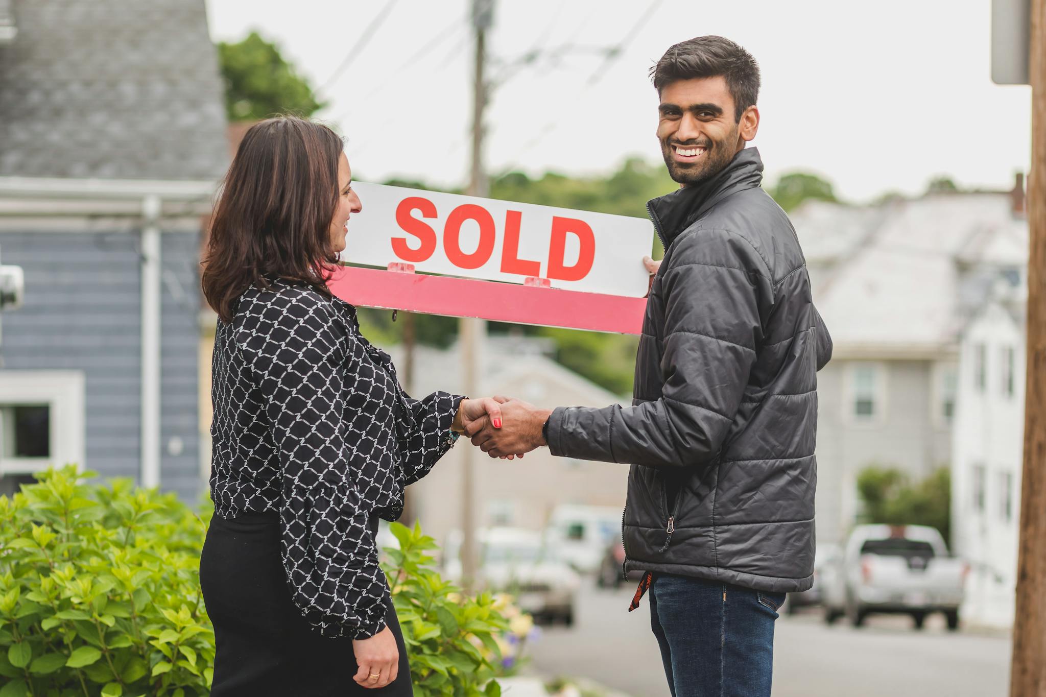 Couple shakes hands in front of new property holding sold sign, symbolizing new home ownership.