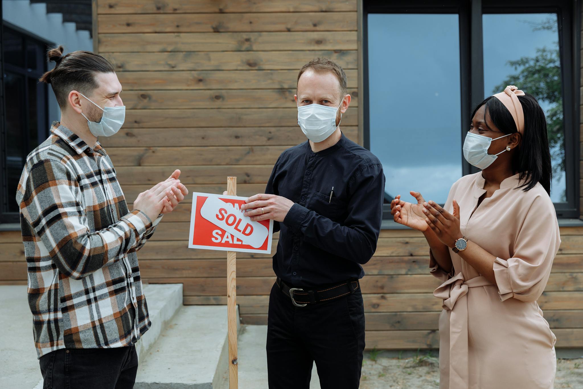 Real estate agent and clients celebrate sale with masks and a sold sign outdoors.