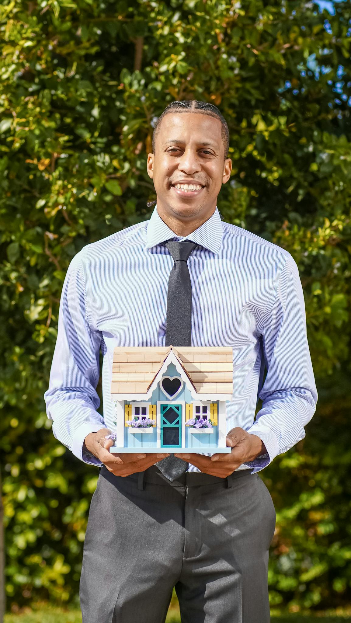 Smiling real estate agent holding a model house in an outdoor setting.
